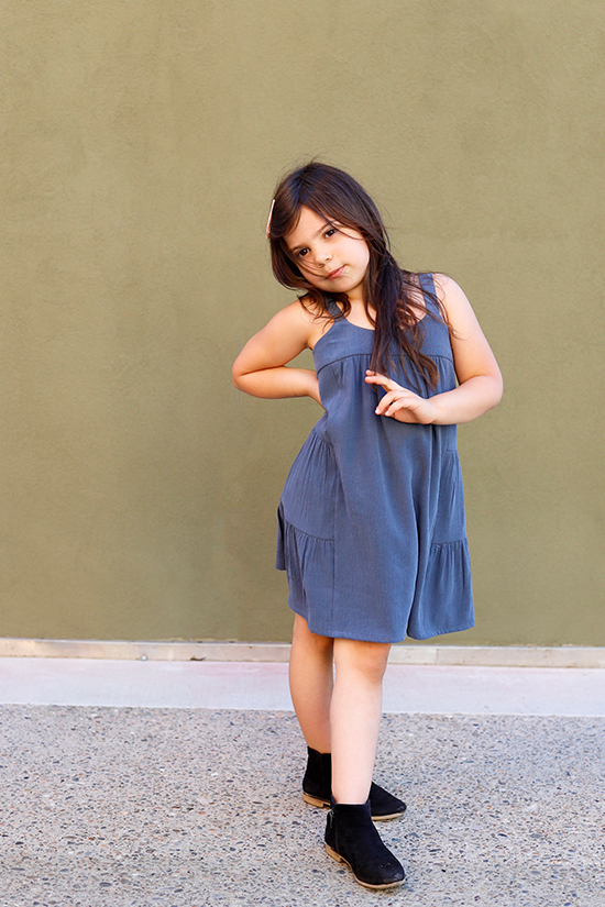 Child stands in front of green wall wearing a blue mini length Mini Marcel Dress