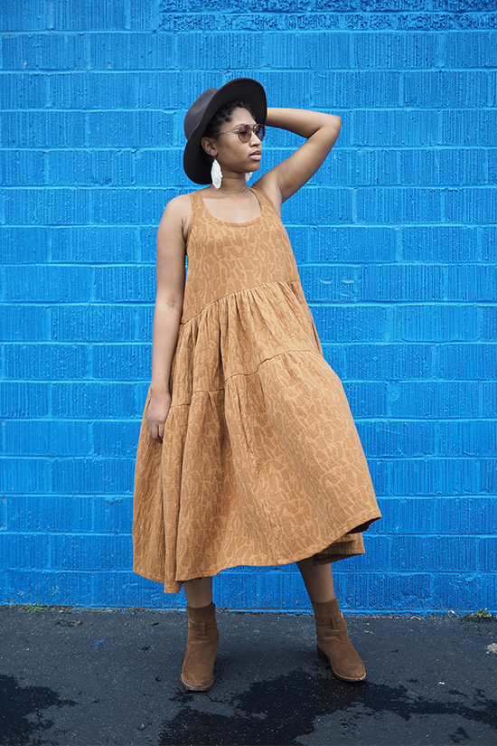 Woman standing in front of blue wall has hand on head and is wearing a caramel colored tiered dress which is blowing in the wind. 