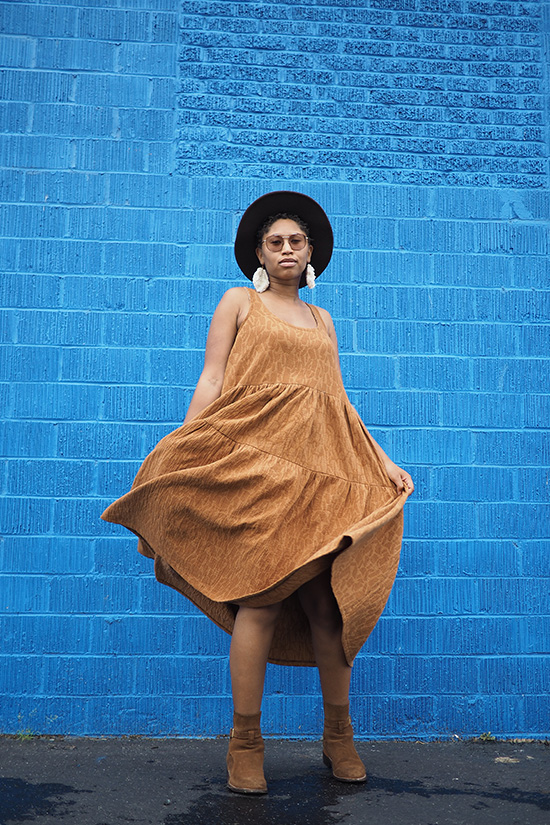Woman wearing a caramel colored tiered dress twirls in front of a blue wall.