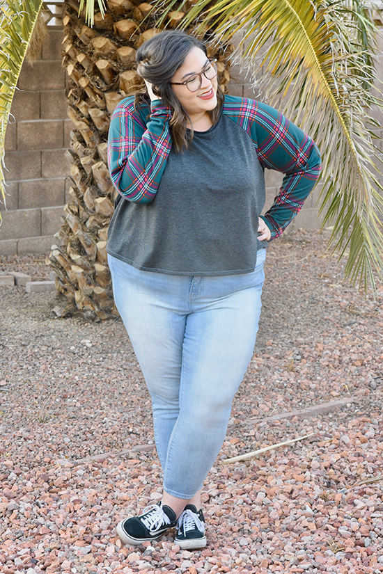 Woman wearing cropped Waterfall Raglan with grey bodice and plaid sleeves stands in front of tree and fence. 