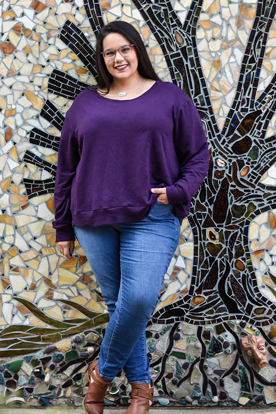 A woman wearing a purple long sleeved Pixie Tee stands in front of a mosaic wall. 