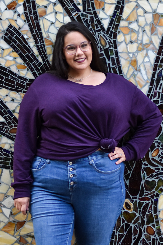 Woman in tied long sleeve purple pixie tee stands in front of mosaic wall. 