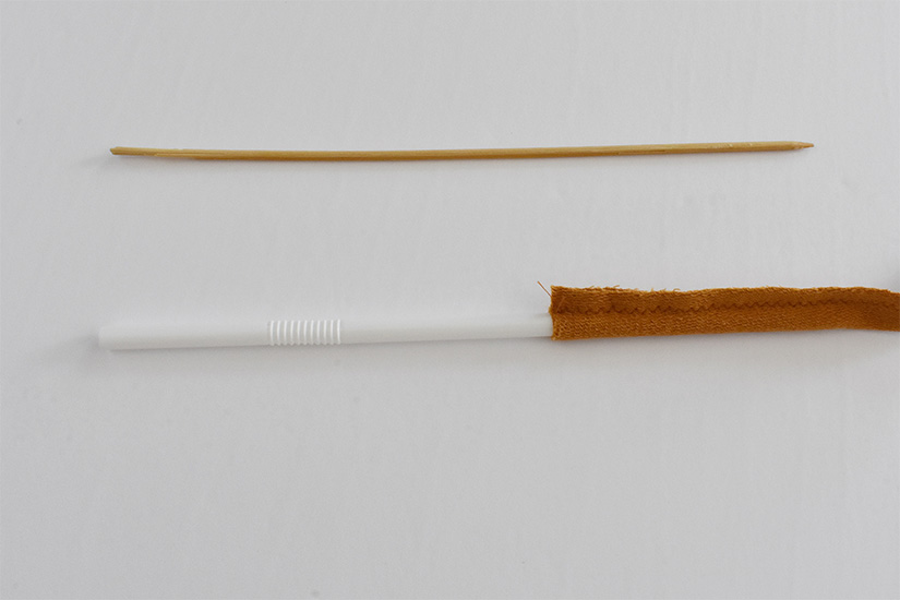 A straw is inserted into a tube of yellow knit fabric. A wooden skewer sits on the white background.