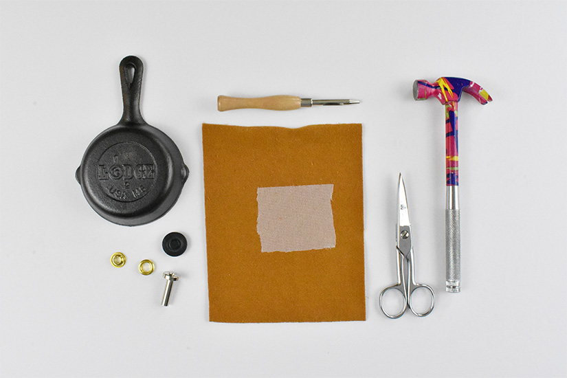 A small cast iron skillet, grommets, an awl, yellow fabric, interfacing, scissors, and a small hammer are arranged on a white background