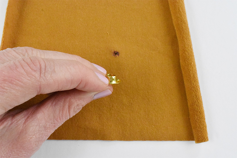 A brass colored grommet piece is held above a small hole cut into a yellow knit fabric 