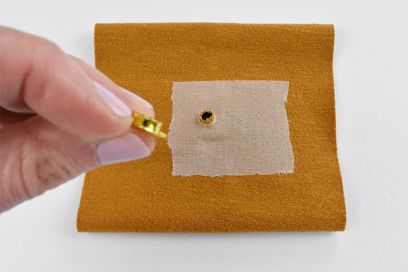 The wrong side of a yellow knit fabric is shown with the larger part of a grommet inserted into a small hole. The smaller part of the grommet is held above the fabric. 