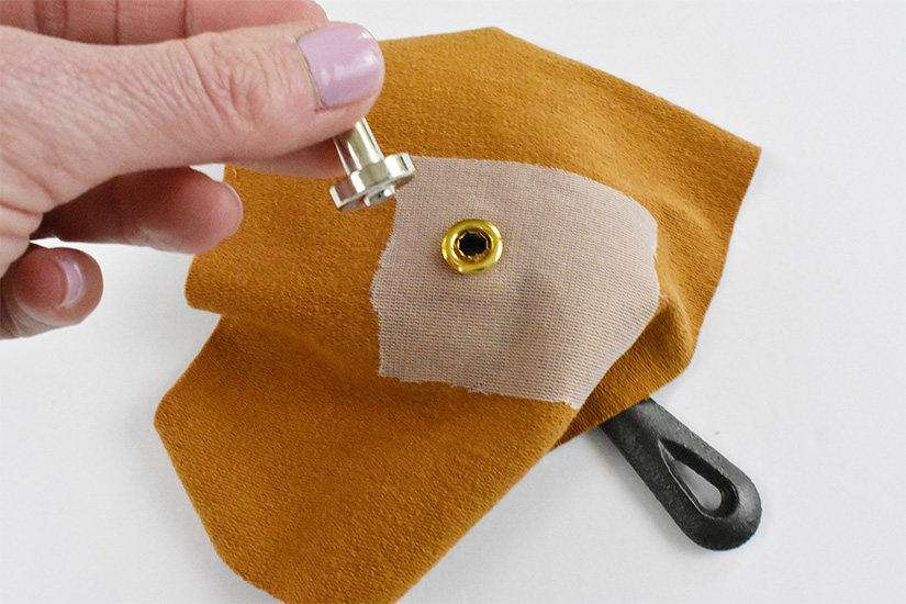 A silver setting tool is held above the back of a brass grommet inserted into yellow knit fabric. The handle of a cast iron skillet can be seen peeking out from under the fabric.