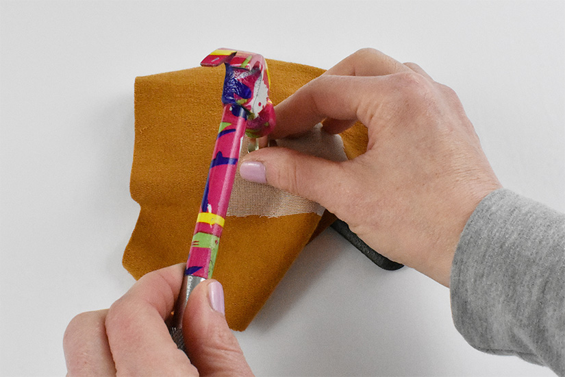 Hands hold a hammer as it hits the top of a silver setting tool placed on the grommet inserted into yellow knit fabric.