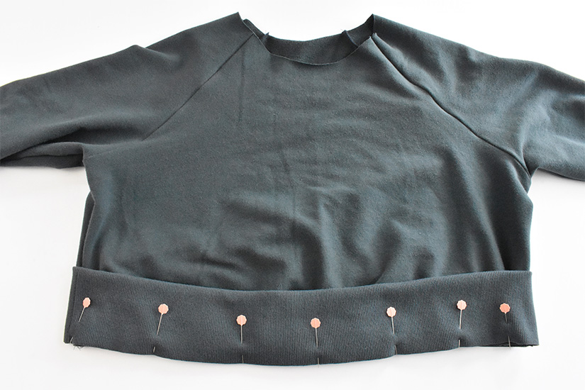 Rib knit hem band in blue/green fabric is shown pinned to bottom of hoodie bodice. 