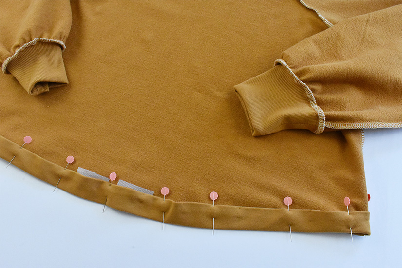 The hem of a gold colored hoodie is shown pinned and pressed to the wrong side by 1"