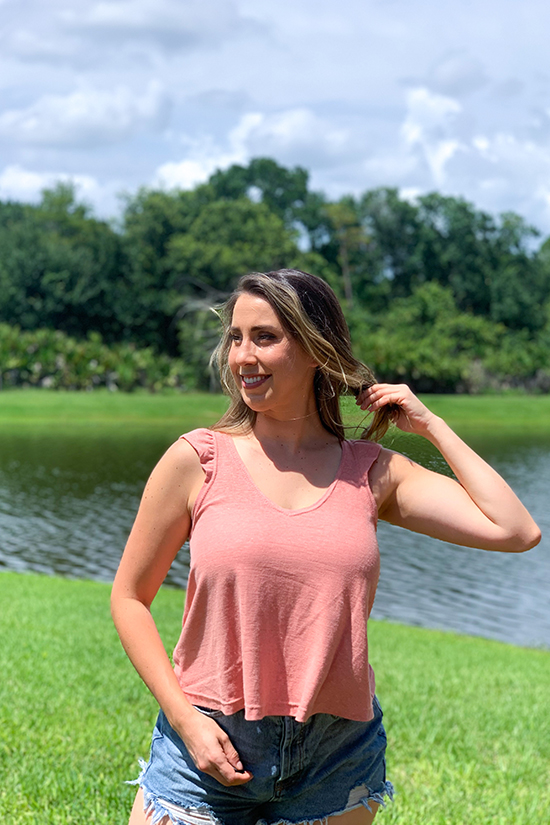 Olivia stands in front of a pond and wears a pink ruffle band tank top with jean shorts. 