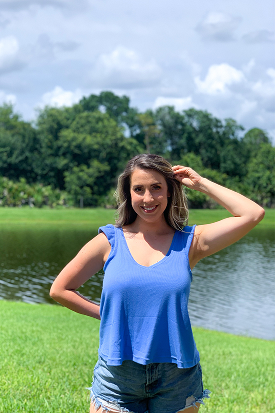Olivia stands in front of a pond and wears a blue ruffle band tank top with jean shorts. 