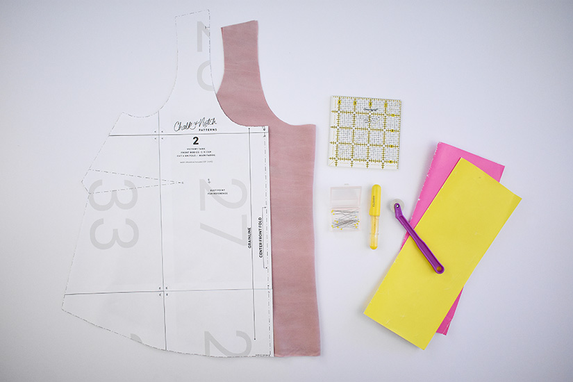 A paper bodice sewing pattern, fabric and assorted supplies for marking and sewing darts lay on a white background. 