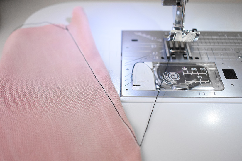 A dart is shown stitched on pink fabric with a long black thread tail still attached to the machine. 