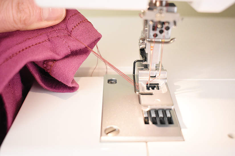 A completed sewn hem is being pulled away from the foot of a coverstitch machine.