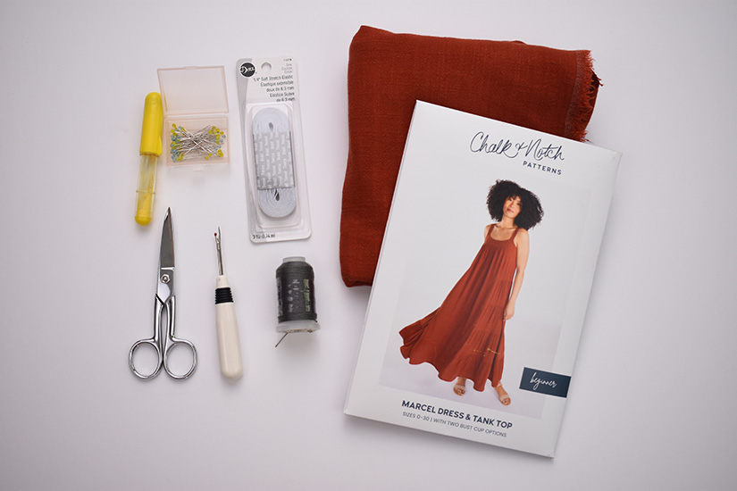 A Marcel dress sewing pattern, orange fabric, and various sewing supplies are shown on a white background. 